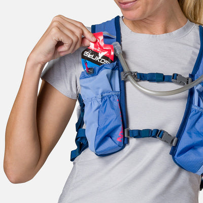 color:Periwinkle/Estate Blue||seo:Nathan TrailMix 2.0 12 Liter Unisex Hydration Pack - Periwinkle/Estate Blue - Female Runner Pulling Glukos Energy Gel (Fruit Punch Flavor) From Front Storage Pocket