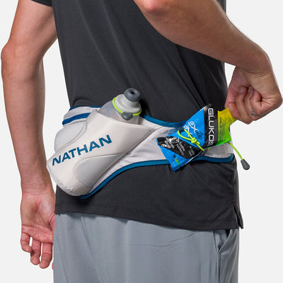 color:Vapor Grey/Sailor Blue||seo:Nathan Peak Hydration Waist Pack - Vapor Grey/Sailor Blue - On Model -  Detail View of Elastic Keepers Holding Glukos Energy Gel (Lemon Lime Flavor)